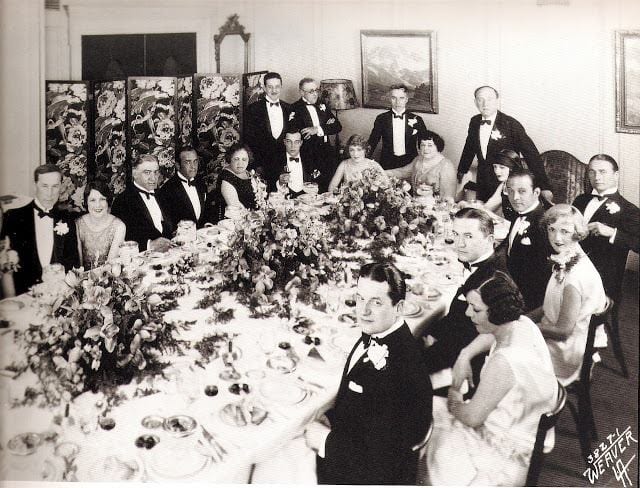 An old style photo of many men and women sitting at a table together ready to eat a meal